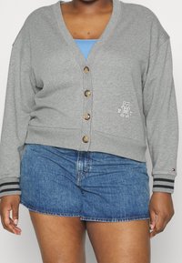 Szary kardigan wykonany z miękkiego materiału, z dekoltem w serek, pięcioma guzikami, wyszywanym logo oraz paskowanymi mankietami. W połączeniu z dżinsowymi szortami.