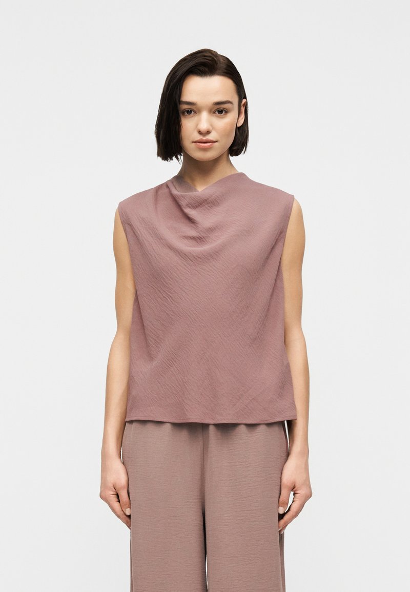 Γυναίκα με κοντά σκούρα μαλλιά που φορά ανδρική αμάνικη μπλούζα σε υφασμάτινη αποχρώση μωβ-ροδακινί και αντίστοιχο παντελόνι με φαρδιά πόδια σε απλό φωτεινό φόντο.