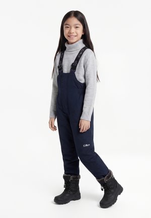 Κορίτσι που φοράει γκρι πουλόβερ με ζιβάγκο, ναυτικά αδιάβροχα παντελόνια με τιράντες και μαύρα χειμερινά μποτάκια με γούνα, χαμογελαστή και όρθια.