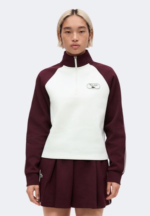 Persoon draagt een witte en donkerrode sweater met een kwart-rits, een logopatch en een bijpassende donkerrode geplooide rok.