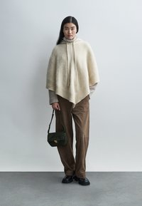 Μπέζ μάλλινο πόνο με κουκούλα και κορδόνια, φορεμένο πάνω από ένα υφασμάτινο ανοιχτόχρωμο πουλόβερ, συνδυασμένο με καφέ παντελόνι κοτλέ και πράσινη τσάντα.