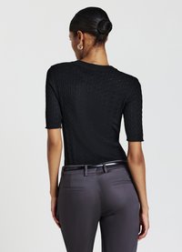 Schwarzes kurzärmeliges Strickoberteil mit strukturiertem Kabeldesign, kombiniert mit grauen taillierten Hosen, die einen eleganten Bund und zwei Gesäßtaschen aufweisen.