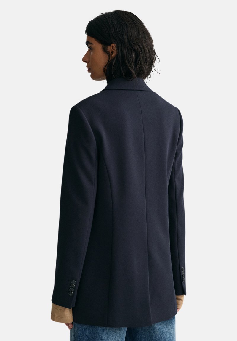 Frau mit kurzem dunklen Haar, die ein navyblaues Blazer über beigen Ärmeln und einer blauen Jeans trägt, blickt nach links vor einem schlichten Hintergrund.