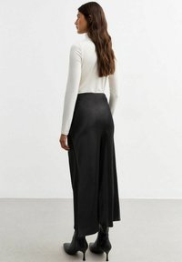 Haut à manches longues blanc associé à une jupe en satin noir, avec une texture lisse, un design fluide et un détail de fente haute, portée avec des bottes à talons noires.