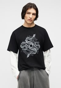 Νεαρό άτομο με σκούρα μαλλιά μέχρι τους ώμους, φορώντας μαύρο T-shirt με λευκό γραφικό φιδιού και λευκά μακριά μανίκια σε στρώσεις, γκρι παντελόνι.