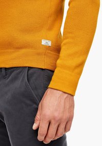 Senfgelbes Strickpullover mit strukturiertem Stoff, ausgestattet mit einem Etikett an der Seite und einem figurbetonten Design. Kombiniert mit dunkelgrauen Hosen.