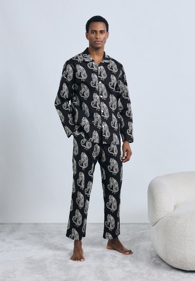 Set di pigiami neri con grandi motivi di tigre bianca. Camicia con bottoni, colletto e maniche lunghe; pantaloni abbinati a taglio largo. Piedi nudi.