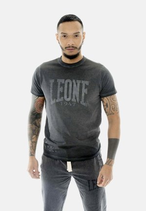 Man met kort haar draagt een donkergrijs Leone 1947 T-shirt en bijpassende korte broek, toont volledige armtattoos, staande tegen een effen achtergrond.