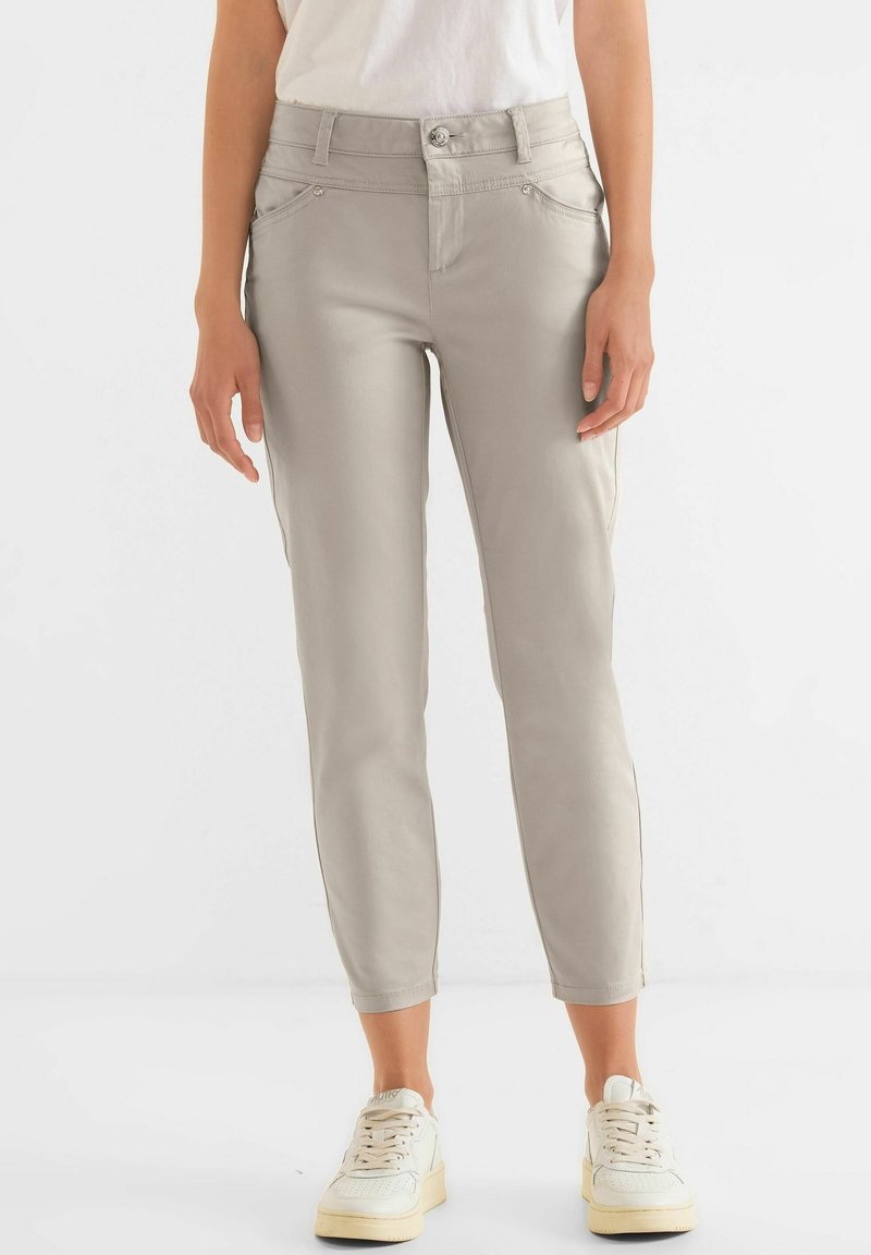 Femme portant un pantalon slim gris clair coupé à la cheville avec un bouton et une fermeture éclair, assorti à des baskets blanches et un haut blanc.