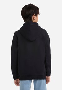 Jugendlicher, der sich abkehrt und einen schwarzen Kapuzenpullover mit herabgezogenem Kapuzen und hellblauen Jeans trägt, vor einem schlichten weißen Hintergrund.