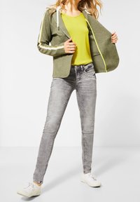 Zelená kapucová bunda s bílými pruhy, žlutý top, šedé úzké džíny a bílé tenisky. Zaměření na volný vrstvený styl a barevné kombinace.