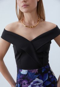 Svart avklädd topp med åtsittande design, med en inneslutande effekt. Buren med ett gyllene, texturerat statementhalsband. Kjol med blommönster.