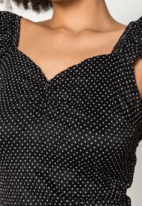 Schwarzes Top mit Punkten, V-Ausschnitt, strukturiertem Stoff und Rüschenträgern. Besitzt geraffte Details an der Brust.
