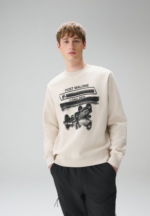 Junger Mann trägt einen beigen Pullover mit schwarzem Schriftzug "POST MALONE LONG BED" und einer kopfüber stehenden Auto-Grafik, kombiniert mit schwarzen Hosen.