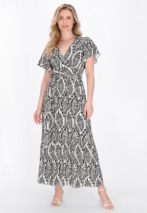 Vrouw staand, gekleed in een maxi-jurk met zwart-witte bladprint, met wijd uitlopende mouwen en openhielsandalen, op een witte achtergrond.