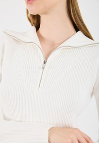 Prim-plan cu o femeie îmbrăcată �într-un pulover alb din tricot cu nervuri, cu un fermoar argintiu parțial deschis la guler.