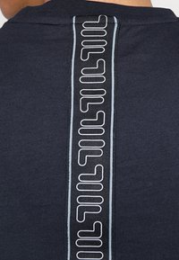 Marinblå T-shirt med ett vertikalt, texturerat svart streck med vit grafisk text längs mitten på baksidan. Mjuk tyg med en slät textur.