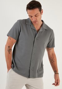 Camisa de botones gris de manga corta, con un patrón texturizado, cuello con muesca y sutiles rayas verticales; cuenta con botones en el frente y un corte relajado.