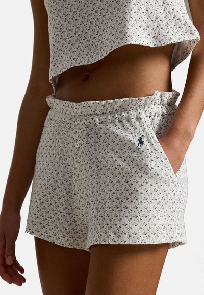 Femme portant un short blanc à fleurs avec des boutons et une taille élastique, assorti d'un haut court coordonné, main dans la poche.