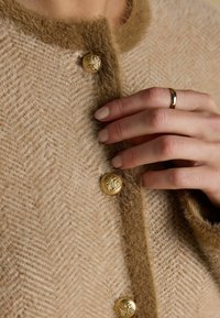 Beež herringbone-stiilis kardigan pehme ja karvase pruuni kraega. Omab kuldseid nuppe, millel on pitsiline disain. Käsi on nähtavalt nuppude lähedal puhkamas.