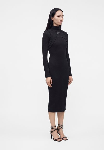 Femme debout portant une robe midi noire moulante à manches longues avec le logo "OFF" et des sandales à talons hauts noires à brides sur fond blanc.