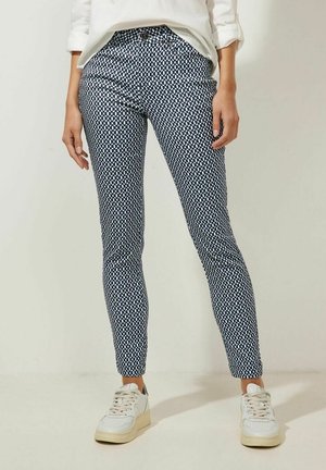 Persoon draagt nauwsluitende marineblauwe en witte broek met geometrisch patroon en witte sneakers, staande tegen een effen lichte achtergrond.