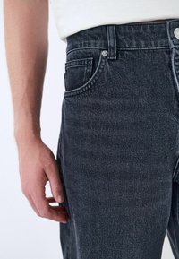 Primer plano de una persona que lleva vaqueros de cintura alta negros con un bolsillo frontal y una camiseta blanca, con la mano relajada a su lado.