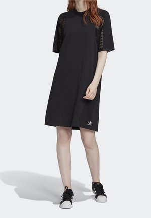 Femme portant une robe noire longueur genoux avec des panneaux en dentelle sur les manches et des baskets Adidas noires avec des bandes blanches et des bouts renforcés blancs.