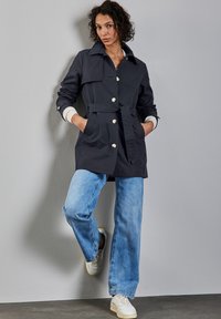 Granatowy płaszcz trench z paskiem w talii, dużymi guzikami z przodu i kontrastowymi, białymi mankietami, w połączeniu z jasnoniebieskimi, szerokimi spodniami dżinsowymi i trampkami.