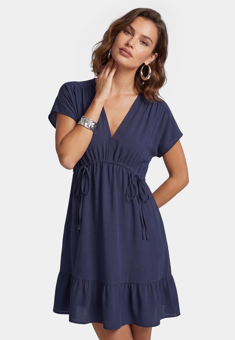Femme portant une robe bleu marine à manches courtes avec un col en V et une taille froncée, accessoirisée avec de grandes boucles d'oreilles créoles et un bracelet en argent.