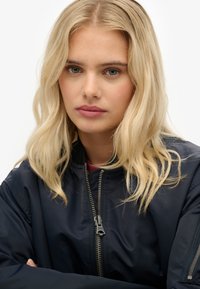 Marine bomberjack met een rits aan de voorkant, geribbelde kraag en een soepele textuur. Model heeft schouderlang blond haar en neutrale make-up.
