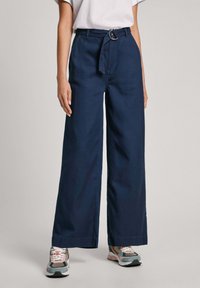 Brede marineblauwe broek met hoge taille, voorzien van een stoffen riem en metalen gesp. Gladde textuur en rechte snit. Draagt met gestreepte sneakers.