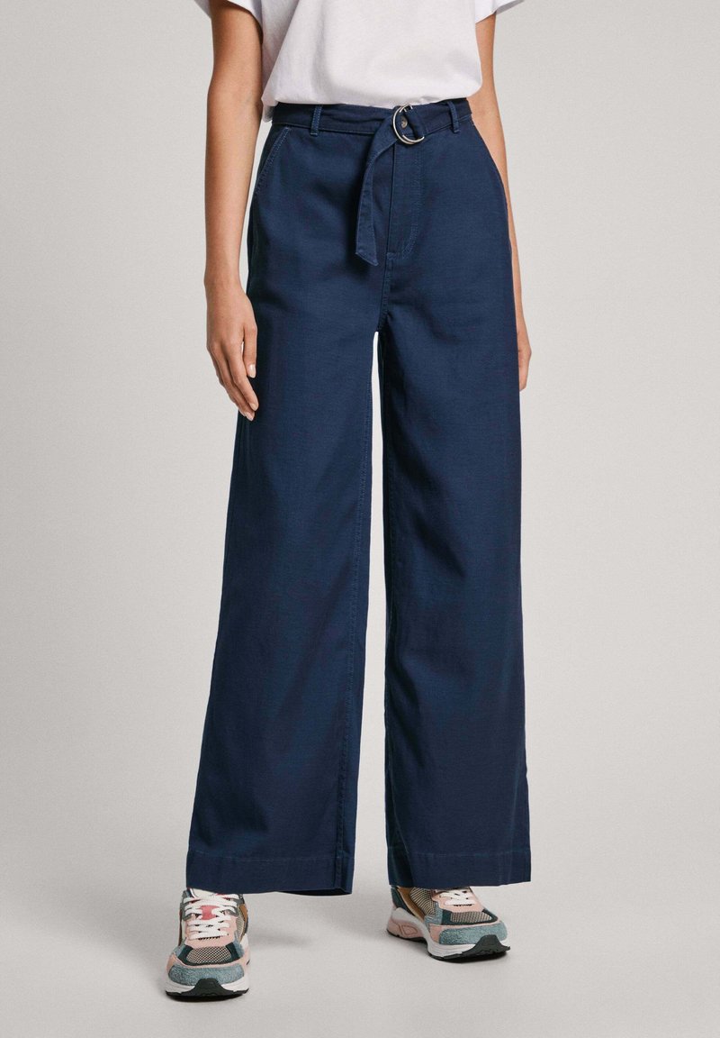 Brede marineblauwe broek met hoge taille, voorzien van een stoffen riem en metalen gesp. Gladde textuur en rechte snit. Draagt met gestreepte sneakers.