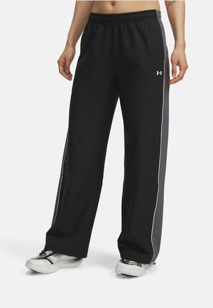 Personne portant un pantalon de jogging noir ample avec des panneaux latéraux gris et des baskets blanches, debout devant un fond uni.