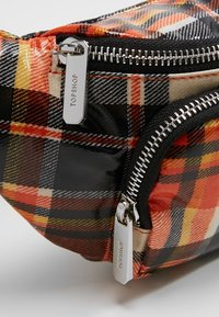 Banane à carreaux noirs et rouges avec finition brillante, dotée de deux fermetures éclair argentées et d'un tire-zippe de marque. Forme arrondie avec texture textile.