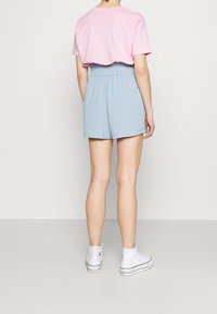 Personne portant un haut à manches courtes rose, un short taille haute bleu clair et des baskets montantes blanches, se tenant devant un fond clair uni.