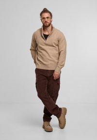 Beige zippered sweater, brown corduroy cargo pants, and tan boots. Model has glasses and a ponytail, standing against a light gray background.