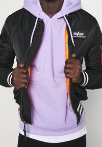 Černá bomber bunda s lesklým povrchem, bílé pruhy na manžetách, oranžové podšívky, spárovaná s levandulovou mikinou. Viditelný detail zipu.
