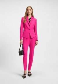 Costume ajusté fuchsia avec une veste cintrée et un pantalon slim, ornés de boutons noirs et de talons noirs élégants. Un sac à main noir apporte un contraste.