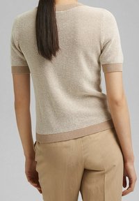 Beżowy sweter na krótki rękaw z dzianiny o teksturowanym splocie, z brązowymi prążkowanymi wykończeniami na dołach i mankietach, noszony z jasnobeżowymi spodniami.