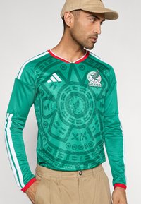 Man som bär en grön Adidas-fotbollströja med långa ärmar, Mexikos logotyp, Aztekiskalenderdesign, beige byxor och en beige korduroymössa som tittar åt höger.