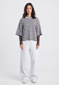 Woman standing facing forward, wearing wide striped shirt over brown lace sleeves, loose white pants, and white sneakers on plain background.