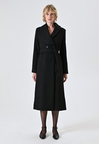 Donna in piedi, che indossa un lungo cappotto nero con cintura e colletto sciallato, collant neri trasparenti e scarpe col tacco a cinturino alla caviglia in pelle verniciata nera.