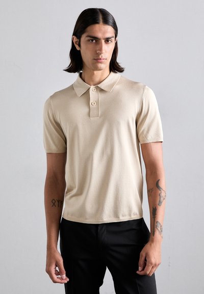 Beige Kurzarm-Polohemd aus weichem Strickmaterial, mit einem klassischen Kragen und einer Knopfleiste mit drei Knöpfen. Schwarze Hosen sind darunter sichtbar.