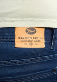 Tummansiniset farkut, joissa on vaaleanruskea nahkaleima. Leimassa on tekstit "Petrol", "Denim Driven Since 1989" sekä ompeluyksityiskohtia.