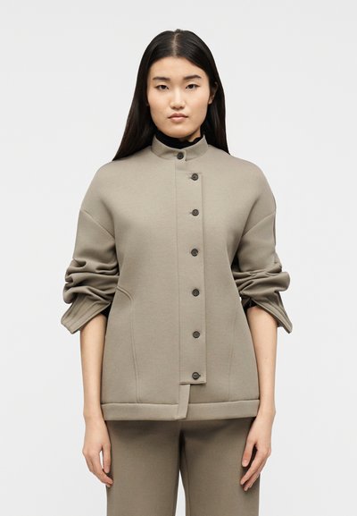 Femme aux longs cheveux noirs portant une veste beige boutonnée à manches froncées sur un pantalon assorti, debout devant un fond clair uni.
