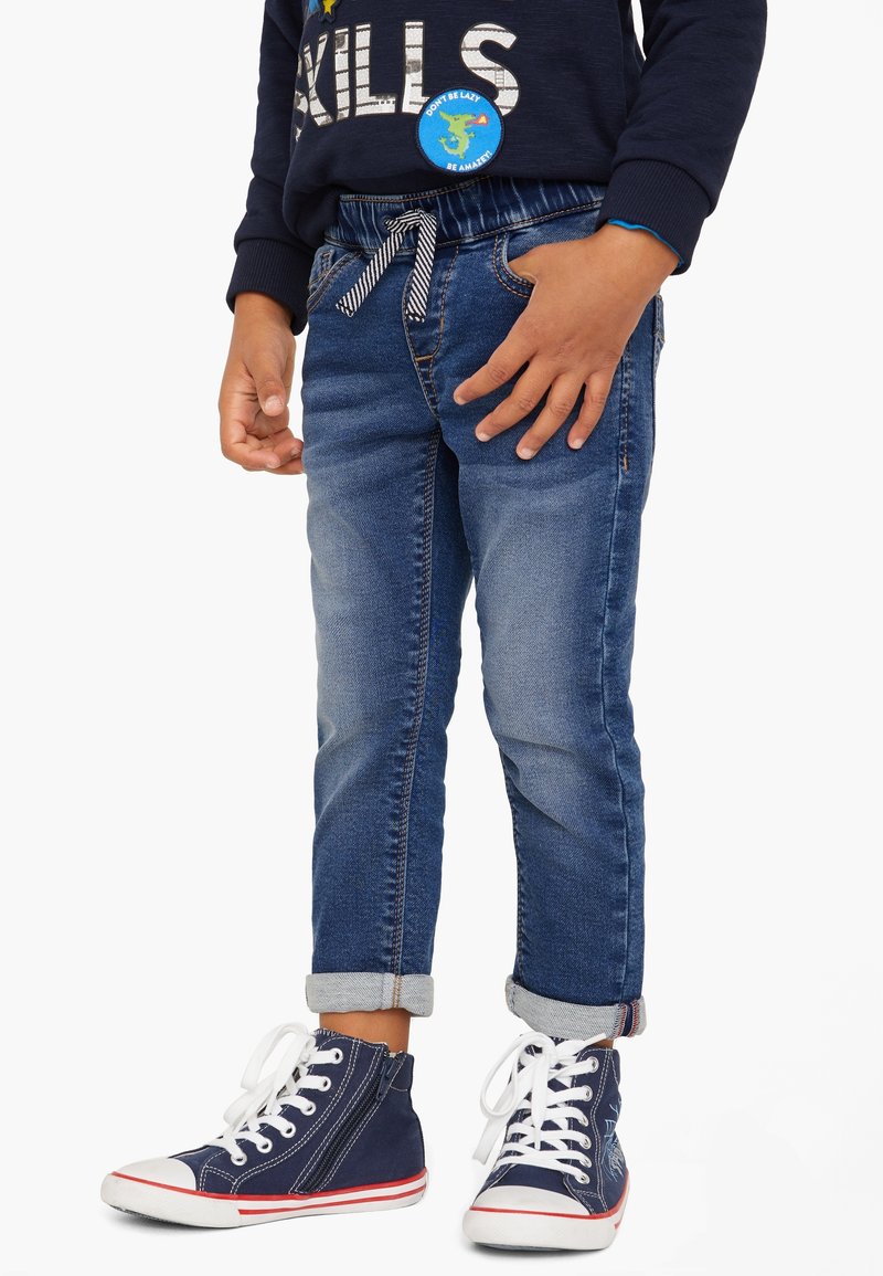 Jean en denim avec une coupe fuselée, de couleur bleue, taille élastique et revers roulés, associé à des baskets montantes bleu marine à lacets blancs.