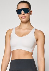 Bela športna modrc z gladko teksturo, širokimi naramnicami in diskretnim logotipom v koralni barvi. V kombinaciji s črnimi sončnimi očali.