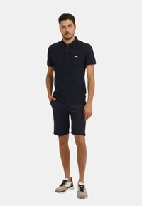 Polo shirt navy con maniche corte, con logo verde. Abbinato a pantaloni corti scuri e sneakers multicolori con suola testurizzata.