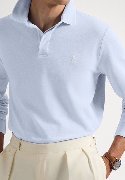 Άνδρας που φοράει ανοιχτό μπλε πουκάμισο πόλο με μακριά μανίκια, μαζεμένο μέσα σε κρεμ πλισέ παντελόνι, με ένα καφέ δερμάτινο ρολόι στο αριστερό καρπό.